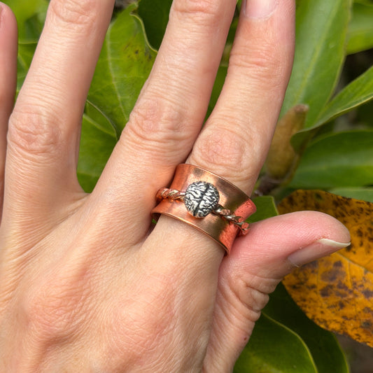 A close-up of a hand wearing a copper ring with a central silver brain, set against a background of green leaves.